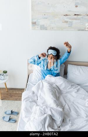 vista ad alto angolo della donna afro-americana compiaciuta nella maschera del sonno che si allunga nel letto Foto Stock