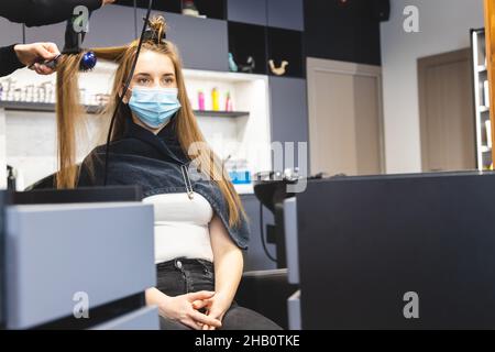 Master donna parrucchiere in una maschera medica asciuga i capelli della ragazza con un asciugacapelli e pettini dopo il lavaggio in un salone di bellezza. Covid-19 pandemia e. Foto Stock