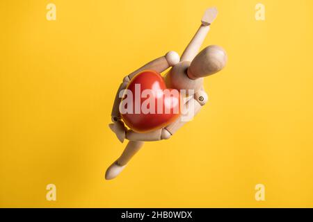 La figurina di legno corre, abbracciando un grande pomodoro rosso sotto forma di un cuore, un simbolo di amore. Sfondo giallo. Messaggio romantico. Foto Stock