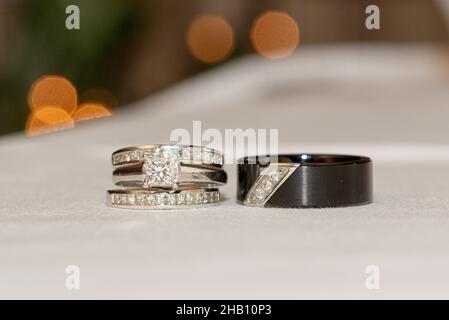 Oro bianco e diamante quadrato sposa anelli di nozze e governa anello nero con diamanti su superficie bianca con bokeh Foto Stock