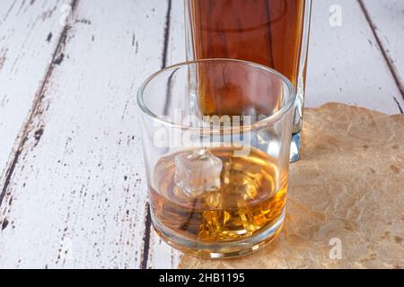 Bicchiere di whiskey con ghiaccio su carta marrone. Sullo sfondo, una bottiglia di whiskey. Foto Stock