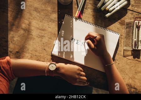 La freelance femminile disegnava la parola 'DESIGN' mentre si siede alla sua postazione di lavoro. Vista ad angolo di una grafica femminile che lavora a un nuovo progetto. Foto Stock