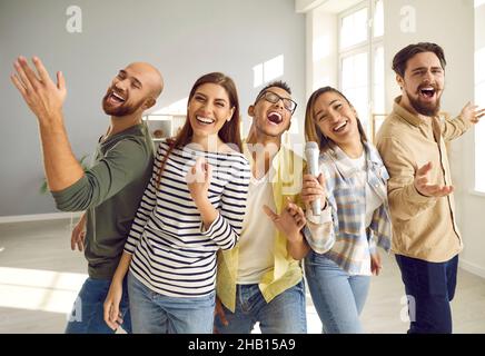 Giovani amici allegri con microfono cantano a voce alta e ridono mentre suonano il karaoke a casa. Foto Stock