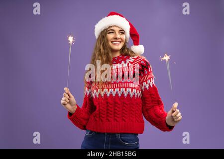 Sorridente giovane donna in balli cappello di Babbo Natale con scintille brucianti su sfondo viola studio Foto Stock