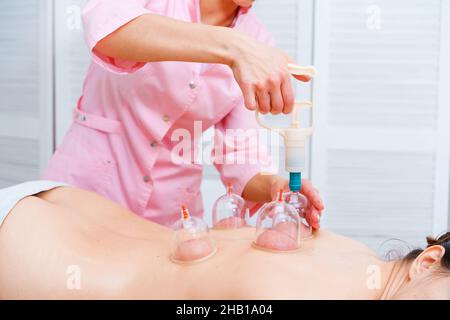 Giovane donna rilassata che riceve inscatolamento sulla schiena a spa. Massaggio sottovuoto. Donna sdraiata sul petto con lattine sulla schiena. Procedura medica. Medico professionista. Foto Stock