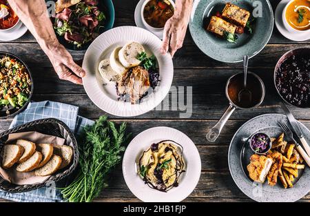 Lo chef raccoglie a mano un piatto con pasto da una vista dall'alto di un tavolo pieno di pasti assortiti, tra cui zuppa, pane, insalata, gnocchi, schnizel, pasta la Foto Stock