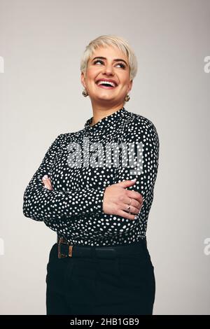 Donna che ride con sicurezza mentre si alza da sola in uno studio. Donna di metà-adulto alla moda che guarda felice mentre in piedi con le sue braccia incrociate contro un Foto Stock