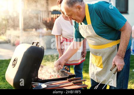 Nonni con grembiuli che grigliano carne e verdure sui bastoni mentre si trova in giardino in primavera. Foto Stock