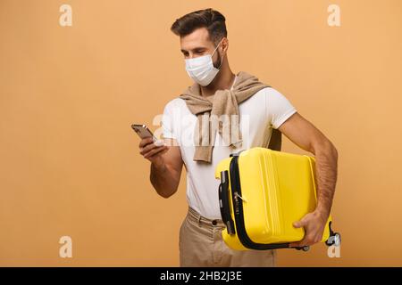 Vita in alto ritratto vista dell'uomo in maschera protettiva che tiene il bagaglio e guardando il suo smartphone mentre controlla i suoi biglietti isolato su sfondo arancione Foto Stock