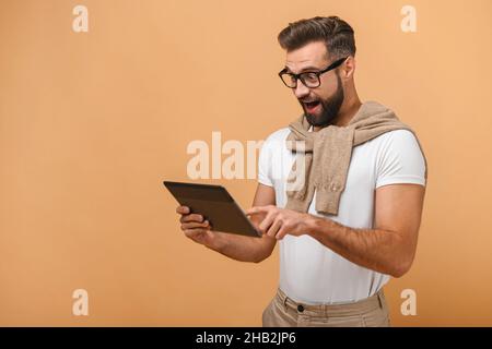 Bell'uomo d'affari con la barba che indossa occhiali che tengono e utilizzano tablet digitale con bocca aperta in sorpresa. Studio girato su sfondo arancione Foto Stock