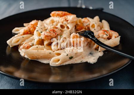 Vista a pezzi di un piatto nero servito con pasta secca penne rigate con una squisita crema di gamberi e gamberi. Foto Stock