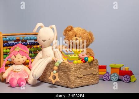 Scatola giocattolo piena di giocattoli per bambini. Contenitore con orsacchiotto, giocattoli in legno soffici ed educativi su sfondo grigio chiaro. Collezione di giocattoli carini per piccoli Foto Stock