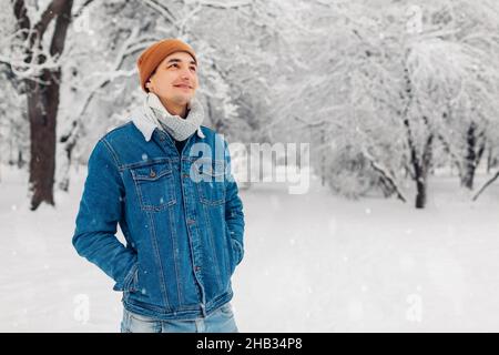 Ritratto di uomo alla moda nel parco nevoso invernale che indossa jeans cappotto a piedi all'aperto. Bel ragazzo ama cadere neve e paesaggio. Moda maschile Foto Stock