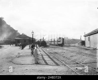 [Stazione ferroviaria di Greymouth], Muir & Moodie studio, studio fotografico, Nuova Zelanda, Stazione ferroviaria di Greymouth preso dal Hokitika fine della piattaforma principale. Sullo sfondo a sinistra è recentemente arrivato un treno alla piattaforma Riverside e i passeggeri sbarcati stanno camminando attraverso l'intersezione di Mackay Street e Mawhera Quay. Sulla destra, sullo sfondo dietro le carrozze, si trovano i vecchi capannoni motore di Greymouth che sono stati demoliti intorno al 1929. Il capannone, in primo piano, fu demolito anche prima del 1969 Foto Stock