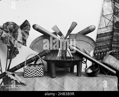 Curios Fijiano, studio Burton Brothers, studio fotografico, circa 1884, Dunedin, Fotografia in bianco e nero, studio allestito di una ciotola di kava, una tazza di kava e un cesto tessuto in cima a un tappeto tessuto, sopra due ventilatori, due pettini per capelli, collane e due club che si incrociano l'uno sull'altro. A sinistra è una pianta tropicale, a destra è una tela di tapa Foto Stock