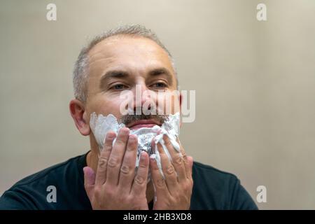 Ritratto di un bell'uomo di mezza età dai capelli grigi che applica schiuma da barba. Affascinante uomo di mezza età rasando la barba e baffi davanti a un Foto Stock