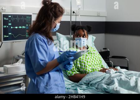 Giovane paziente afroamericano con maschera facciale protettiva contro covid19 sdraiati a letto spiegando sintomi di malattia a infermiere medico in ospedale ufficio. Assistente che controlla il trattamento sanitario Foto Stock
