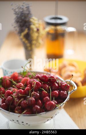 Colazione estiva, ciliegie e fragole in una ciotola, torte con tè, tavolo con fiori. Vista frontale. Foto Stock