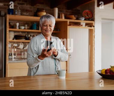 Orientamento ritratto di felice multiculturale anziana femmina scorrimento su smartphone mentre in piedi in cucina moderna. Foto Stock