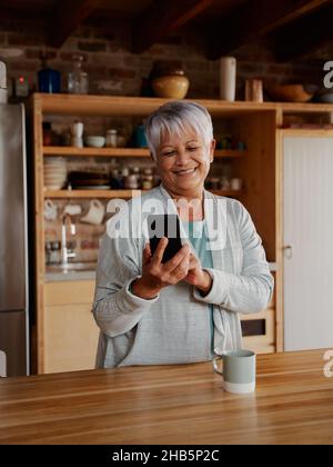 Orientamento ritratto di felice multiculturale anziana femmina digitando un messaggio sullo smartphone mentre si trova in cucina moderna. Foto Stock