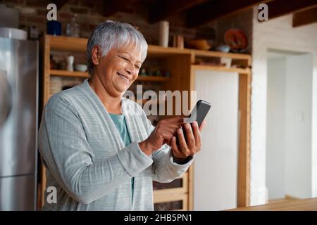 Contenuto multiculturale anziana femmina che scrive un messaggio sullo smartphone mentre si trova in cucina moderna. Foto Stock