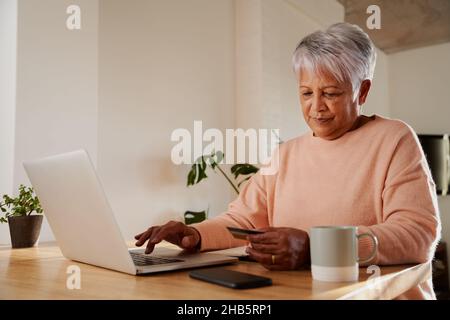 Anziana multi-etnia femminile felicemente digitando nei dettagli bancari per effettuare il pagamento online sul portatile, seduto al banco della cucina. Foto Stock