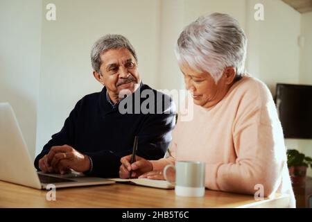 Coppia multiculturale anziana seduta al moderno banco della cucina con computer portatile, moglie che fa appunti in notebook. Budget per il pensionamento. Foto Stock