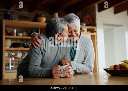 Felicemente anziana coppia biraciale che gode di pensione. Moglie che tiene marito in cucina moderna. Il marito bacia la moglie sulla guancia. Foto Stock