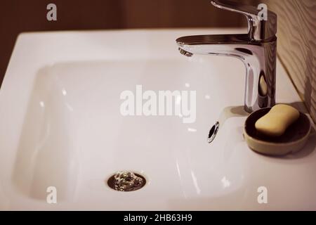 Un lavello vuoto in ceramica bianca, un moderno rubinetto in acciaio inossidabile e accanto un piatto di sapone con sapone bianco. Bagno interno. Pulizia e igiene. Foto Stock