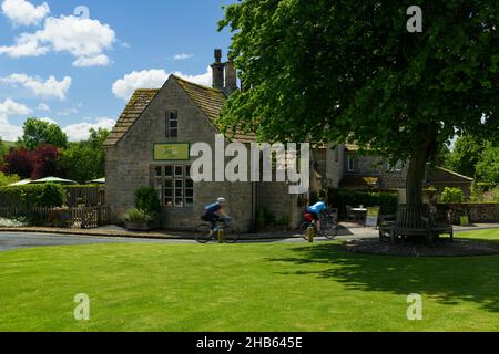 2 ciclisti passando pittoreschi cottage tea rooms caffè & albero sul verde del villaggio, in paesaggio soleggiato rurale Bolton Abbey - Yorkshire Dales, Inghilterra Regno Unito. Foto Stock