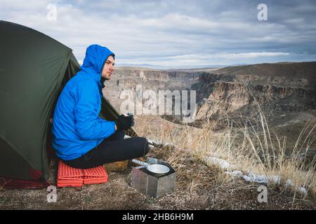 Uomo in blu si siede vicino alla tenda e beve caffè mentre pensa e guarda ai paesaggi della valle di Ihlara. Campeggio nella natura drammatica in T Foto Stock