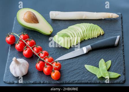 Le mani maschili circa per affettare l'avocado fresco con un coltello da cucina dello chef su un tagliere nero opaco. Cibo sano e concetto di cucina Foto Stock