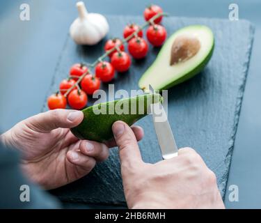 Le mani maschili circa per affettare l'avocado fresco con un coltello da cucina dello chef su un tagliere nero opaco. Cibo sano e concetto di cucina Foto Stock