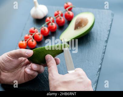 Le mani maschili circa per affettare l'avocado fresco con un coltello da cucina dello chef su un tagliere nero opaco. Cibo sano e concetto di cucina Foto Stock