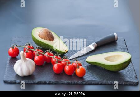 Le mani maschili circa per affettare l'avocado fresco con un coltello da cucina dello chef su un tagliere nero opaco. Cibo sano e concetto di cucina Foto Stock