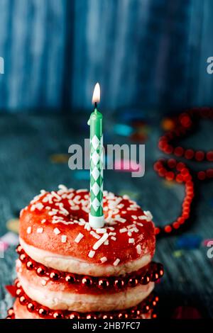 primo piano di una candela illuminata in cima a un mucchio di ciambelle ricoperte di glassa rossa, su un tavolo grigio rustico in legno Foto Stock