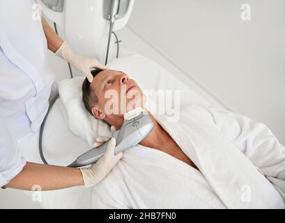 Vista dall'alto della mano estetista che tiene moderno apparato medico ultrasonico e che effettua il sollevamento del viso e ringiovanente trattamento di bellezza anti-invecchiamento su fa Foto Stock