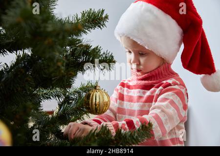 Ragazza in un cappello di Santa appende una palla di Natale su un albero di Natale. Stile di vita. Foto Stock