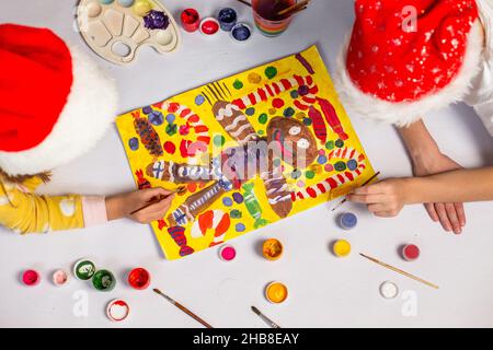 Ragazzo e ragazza in rosso babbo natale cappelli si trovano sul pavimento e disegnare un quadro di Natale a casa. I bambini dipingono un uomo di pan di zenzero con vernici. Foto Stock