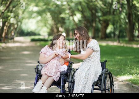 Due donne positive in abiti estivi sorridenti e guardandosi mentre si siedono in carrozzine tra il parco verde. Supporto, amicizia e concetto di persone. Foto Stock