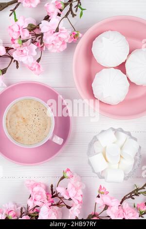 Caffè e marshmallow al mattino per colazione. Fiori di primavera e dessert su sfondo bianco di legno. Disposizione piatta. Carta color rosa pastello per Foto Stock