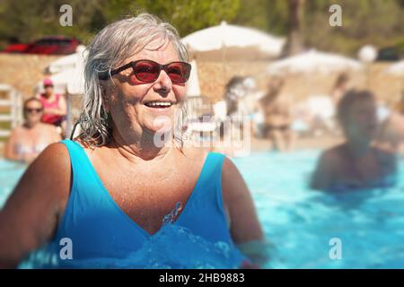 Anziana donna anziana con capelli grigi, con costume da bagno blu che fa aerobica in acqua nella piscina dell'hotel Foto Stock