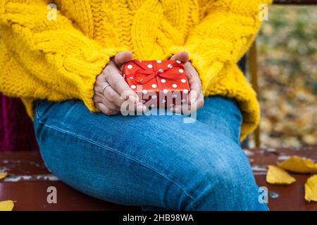 Donna che tiene in mano una scatola regalo rossa. Sorpresa nelle mani femminili. Donna seduta sulla panca del parco Foto Stock