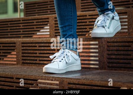 Le gambe femminili indossano jeans e sneakers bianche che camminano sulle scale. Donna con scarpe sportive. Foto Stock