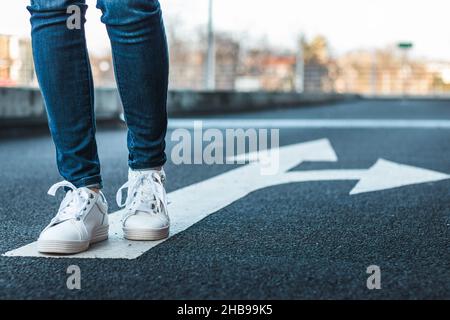 Se si va contro il mainstream potrebbe essere una scelta giusta. Le gambe femminili indossano jeans e scarpe bianche che camminano sul segnale di direzione alla strada. Decisione Foto Stock