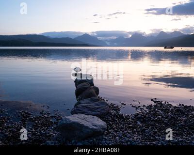 Una linea di pietre che si aggettano nel Lago McDonald nel Parco Nazionale del Ghiacciaio con l'aspra catena montuosa Livingston e una piccola barca a motore nel backgroun Foto Stock