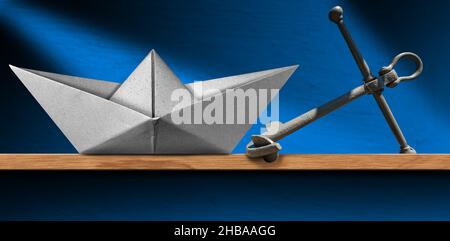 Primo piano di una barca di carta bianca e di un'ancora di metallo grigio su una mensola di legno a casa, con una parete blu sullo sfondo. Foto Stock
