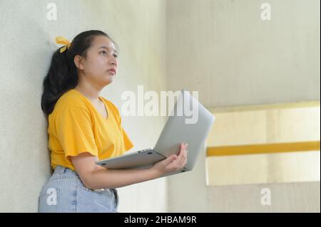 Una donna con un laptop in piedi contro una parete di cemento, indossando una t-shirt bianca e guardando la parte superiore, pensare posa, lavoro a casa, ufficio a casa, remoto Foto Stock