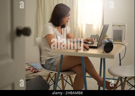 Donna asiatica che si riunisce online mentre lavora a casa, indossando maglietta bianca e occhiali utilizzando un computer portatile sulla scrivania per la conferenza, seduto in camera da letto de Foto Stock