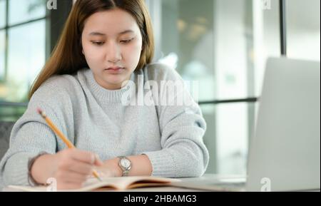 Uno studente adolescente prende appunti con determinazione, indossando un maglione grigio che tiene una matita gialla e un computer portatile sulla scrivania, lavora da casa, studyin Foto Stock